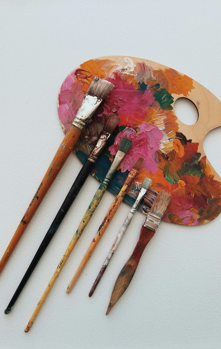 Six used paintbrushes rest on a colorful wooden artist palette against a white background.