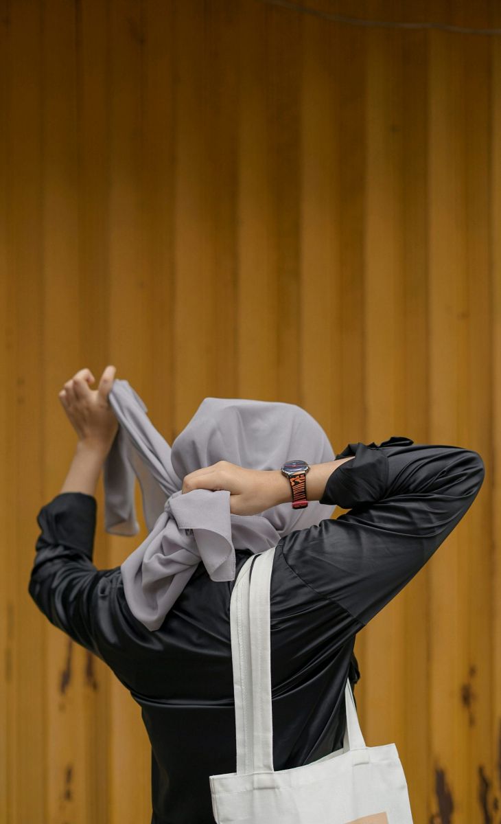 Person in a gray hijab and black shirt adjusts scarf, standing against a yellow corrugated metal wall.