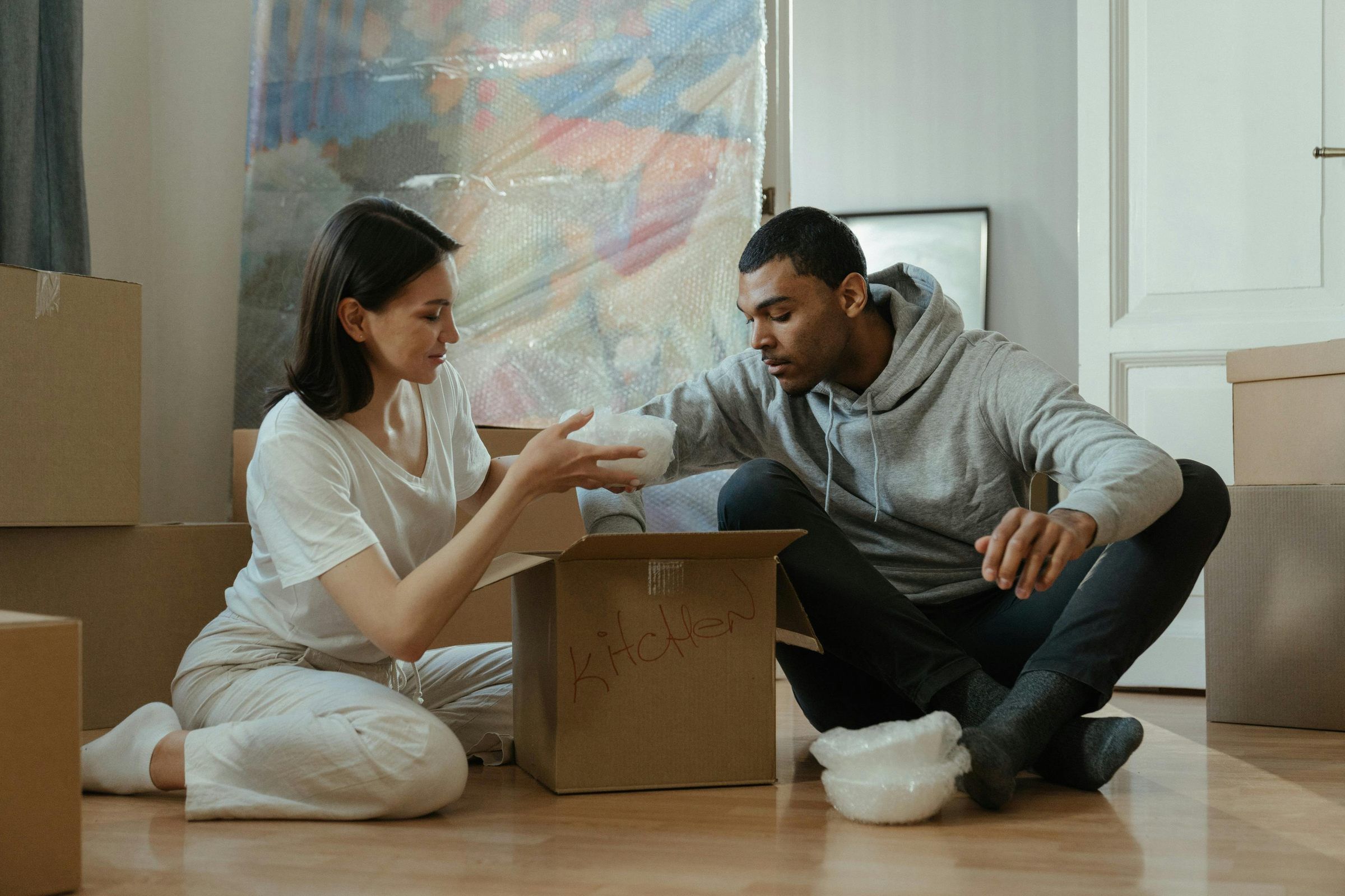 Two people sit on the floor unpacking a box labeled "kitchen" surrounded by moving boxes.