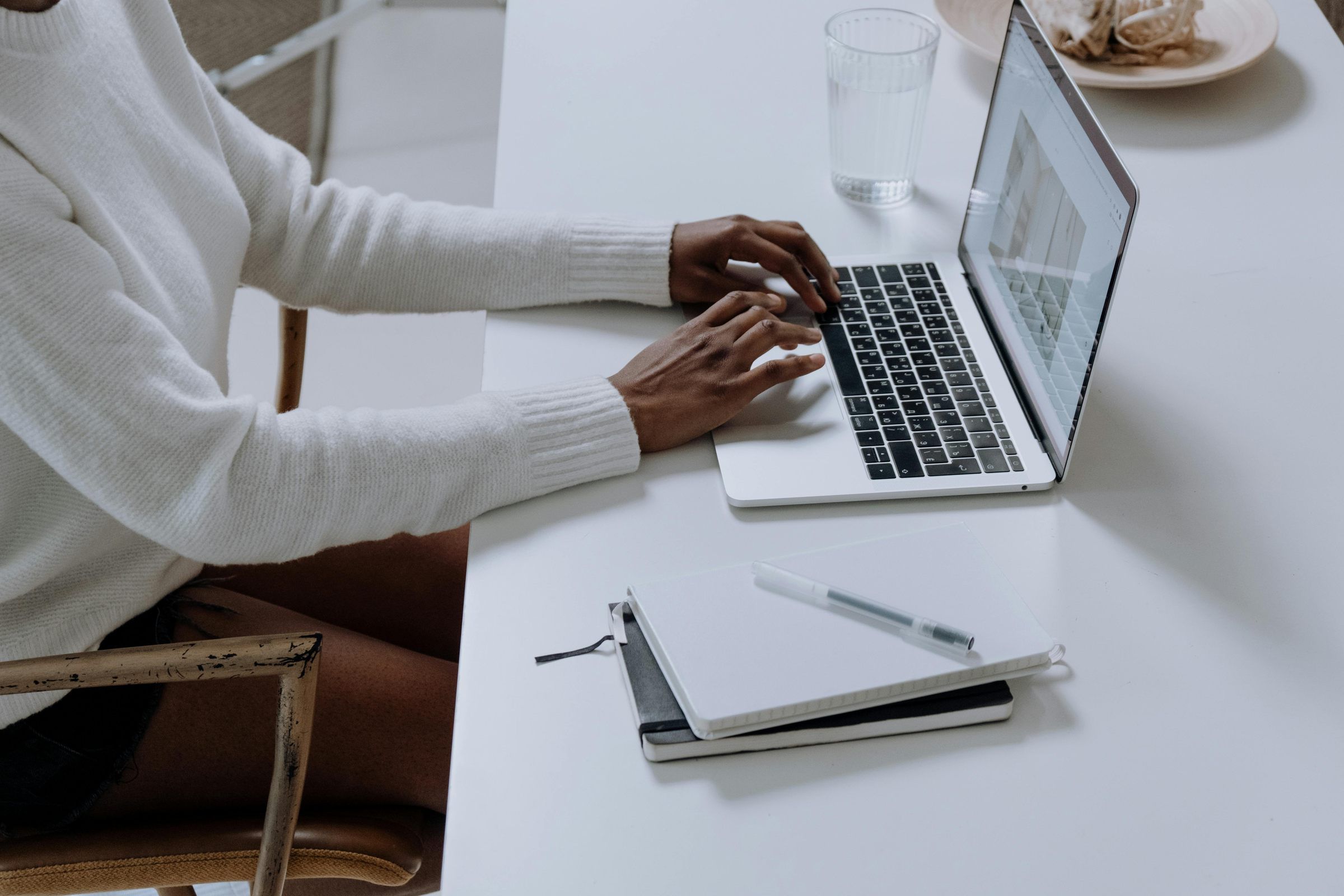 Person typing on a laptop at a white desk with notebooks, a pen, and a glass of water nearby.
