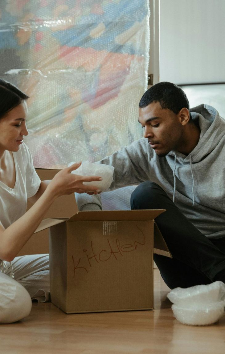 Two people sit on the floor unpacking a box labeled "kitchen" surrounded by moving boxes.