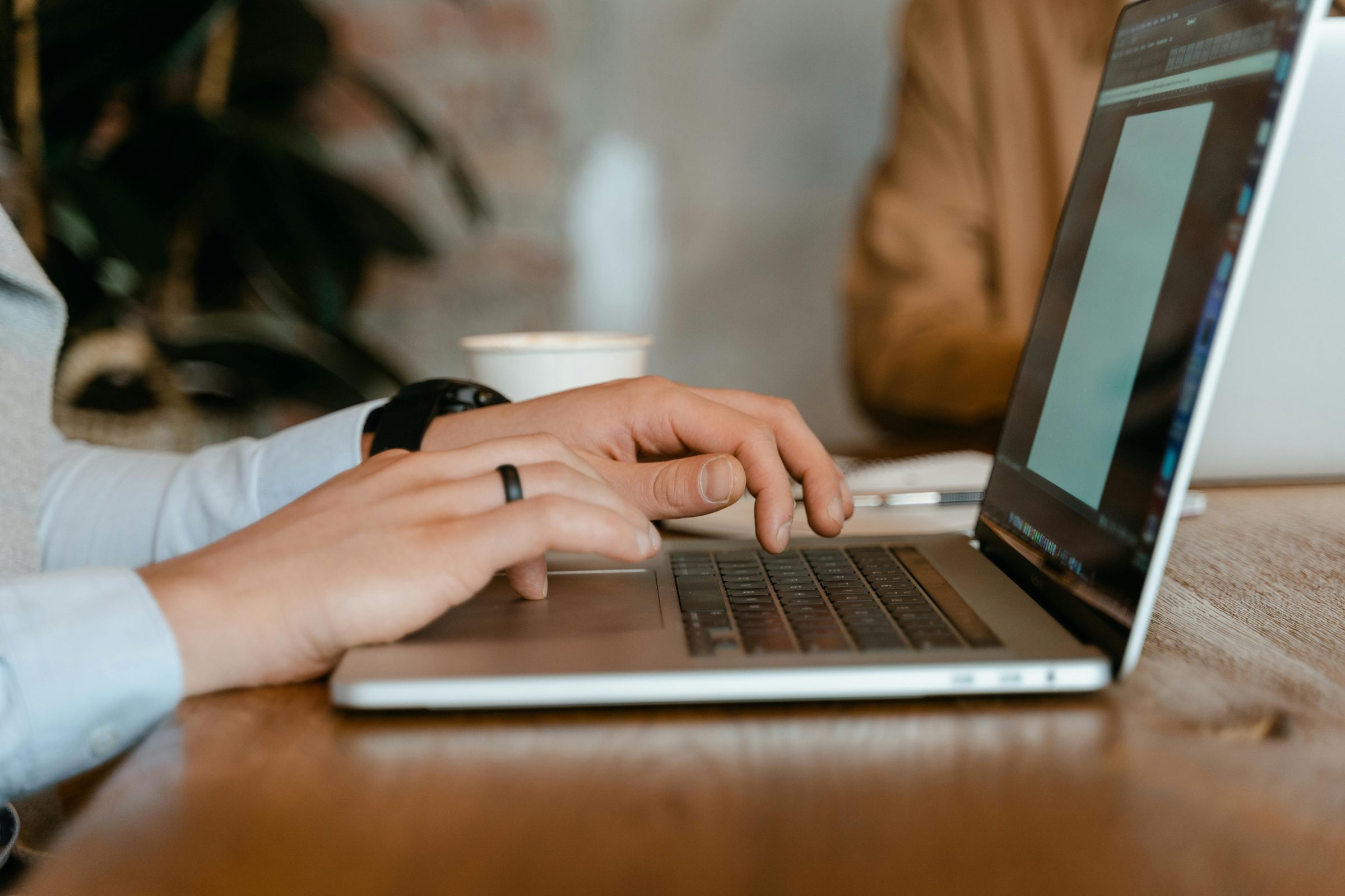 Close-up of hands typing on a laptop keyboard at a wooden table, with a coffee cup in the background.