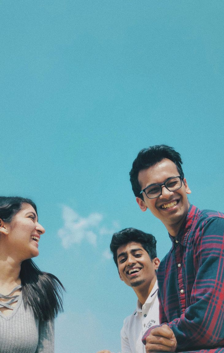 Three young adults smiling and laughing together outdoors against a bright blue sky.